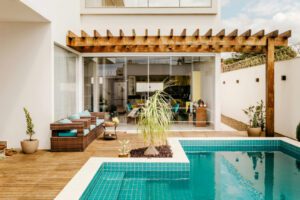 Pictured above is an outdoor deck area with brown, padded chairs underneath a pergola. A blue-tiled swimming pool adjoins the pergola.