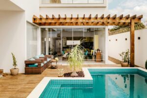 Pictured above is an outdoor deck area with brown, padded chairs underneath a pergola. A blue-tiled swimming pool adjoins the pergola.